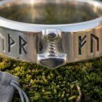 Close up of a polished steel ring with an applied Mjolnir motif between engraved Elder Futhark runes, resting on moss
