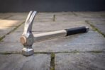 Polished steel rip claw hammer with pale hickory handle, black rubber grip and rune line lying on stone paving