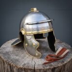 Polished steel Roman helmet front view with brass meander brow band, crest holder and hinged cheek guards on a tree stump