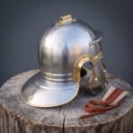 Polished steel Roman helmet with brass edging, neck guard and hinged cheek guard on a tree stump beside leather chin ties
