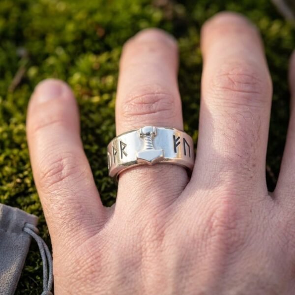 Polished steel rune ring with a raised Mjolnir motif and Elder Futhark band worn on a finger over green moss