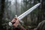Polished steel short sword with knotwork etched fuller, scrolled guard and lobed pommel gripped in a fist in misty woods