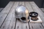 Polished steel spectacle helmet with riveted panels and eye guards on a grey wooden table beside its leather liner and strap