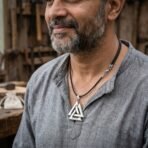 Polished steel valknut pendant with a rune band on a black leather cord, worn by a bearded man in a grey linen shirt in a workshop