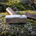 Polished steel war hammer head with runes cut into the face, tan leather wrapped haft, resting on a mossy rock