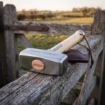 Polished steel war hammer head with cut rune line resting on a weathered gate rail beside a brown leather head cover