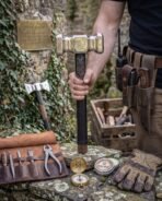 Polished steel war hammer with walnut haft and black leather grip held upright beside a leather tool roll and work gloves