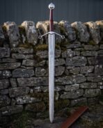 Quatrefoil guard damascus sword with an engraved Valknut panel, oxblood leather grip and steel wheel pommel standing point down against a mossy dry stone wall