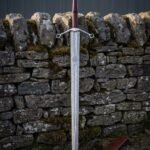 Quatrefoil guard damascus sword with an engraved Valknut panel, oxblood leather grip and steel wheel pommel standing point down against a mossy dry stone wall