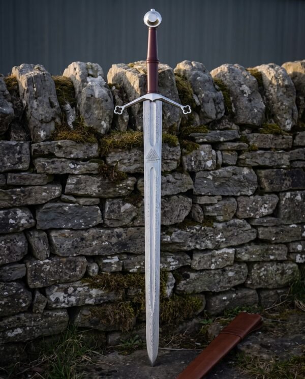 Quatrefoil guard damascus sword with an engraved Valknut panel, oxblood leather grip and steel wheel pommel standing point down against a mossy dry stone wall