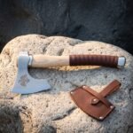 Raven axe with a deep etched Norse bird motif on a hand forged steel bearded head, pale ash haft and leather wrapped grip, resting on a sunlit volcanic boulder
