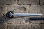Raven basket hilt sword with a blackened cage guard and brass rosette rivets lying on old stone paving beside its black scabbard, blade polished bright.