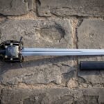 Raven basket hilt sword with a blackened cage guard and brass rosette rivets lying on old stone paving beside its black scabbard, blade polished bright.