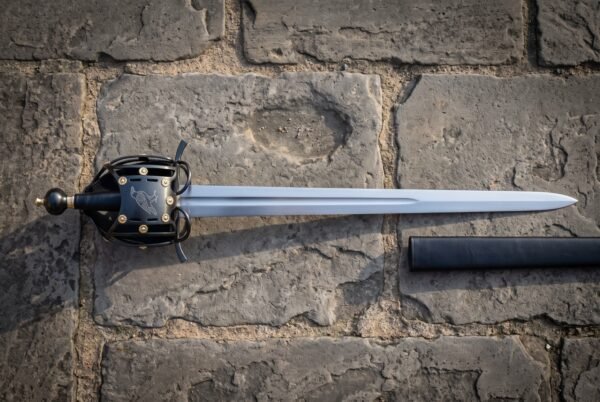 Raven basket hilt sword with a blackened cage guard and brass rosette rivets lying on old stone paving beside its black scabbard, blade polished bright.