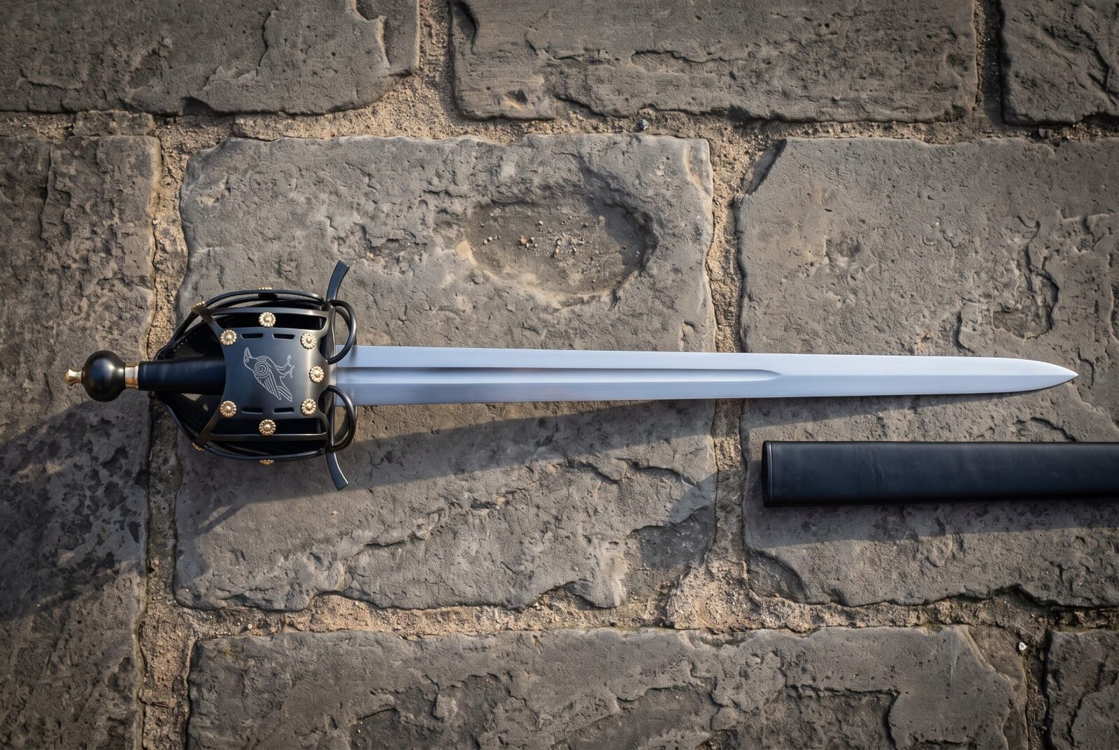 Raven basket hilt sword with a blackened cage guard and brass rosette rivets lying on old stone paving beside its black Raven basket hilt sword with a blackened cage guard and brass rosette rivets lying on old stone paving beside its black scabbard, blade polished bright.