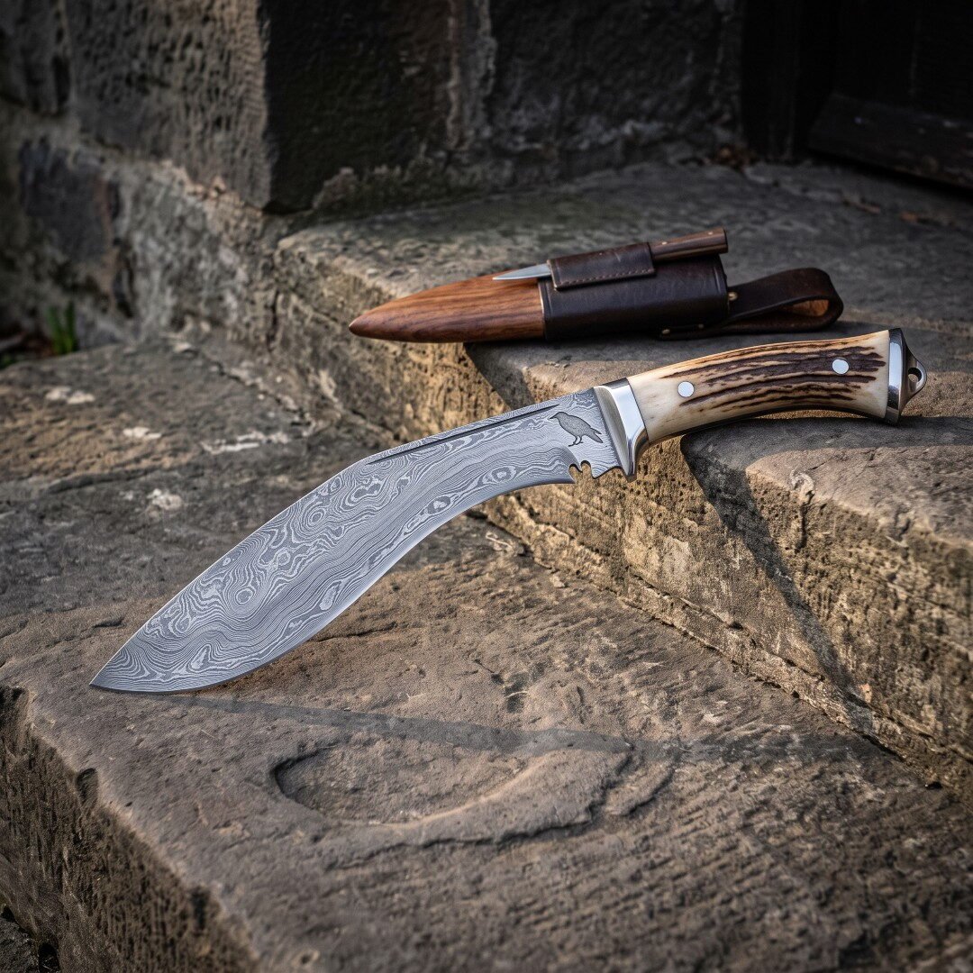 Raven etched damascus kukri with natural stag antler handle and steel bolster resting on a worn stone step beside its le Raven etched damascus kukri with natural stag antler handle and steel bolster resting on a worn stone step beside its leather sheath holding karda and chakmak knives