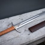 Raven etched falchion in polished 1095 steel lying on a weathered timber bench beside its brown leather scabbard, showing the tan leather grip and faceted pommel.