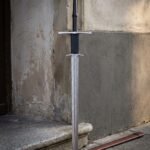 Raven pommel damascus longsword standing against a weathered plaster wall beside a brown leather scabbard, showing the black leather grip and twin steel guards.