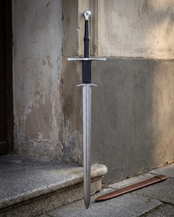 Raven pommel damascus longsword standing against a weathered plaster wall beside a brown leather scabbard, showing the black leather grip and twin steel guards.