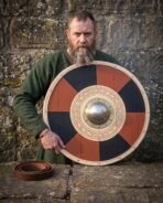 Red and black quartered round shield with brass boss and carved knotwork ring held by a bearded man in a green wool tunic by a stone wall