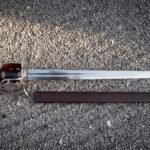 Red lined basket hilt sword in polished 1095 steel lying on pale gravel beside its brown leather scabbard, showing the pierced shell guard and wire bound grip.