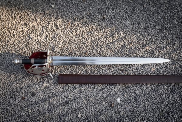 Red lined basket hilt sword in polished 1095 steel lying on pale gravel beside its brown leather scabbard, showing the pierced shell guard and wire bound grip.