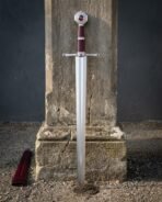 Red stone knightly sword with a mirror polished blade, engraved octagonal pommel and oxblood leather grip standing point down against a cracked stone plinth outdoors