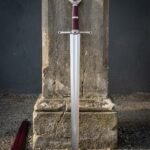 Red stone knightly sword with a mirror polished blade, engraved octagonal pommel and oxblood leather grip standing point down against a cracked stone plinth outdoors
