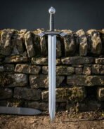 Ring pommel viking sword in 1095 steel with Elder Futhark runes cut across the guard, standing point down against a mossy dry stone wall in low sun