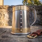 Riveted stainless steel tankard with engraved brass bands top and bottom on a stone step, leather belt hanger at its side