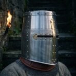 Riveted steel great helm with brass cross and knotwork engraved band worn in profile over a chainmail collar with a burning torch behind
