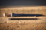 Roman gladius laid on a warm sandstone block beside its dark leather scabbard, blackened ridged blade tapering from a knotwork engraved guard and pale disc pommel
