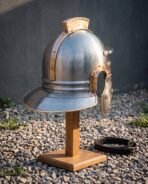 Roman legionary helmet in steel with bronze edging and wide neck guard on a wooden stand over gravel, seen from the side
