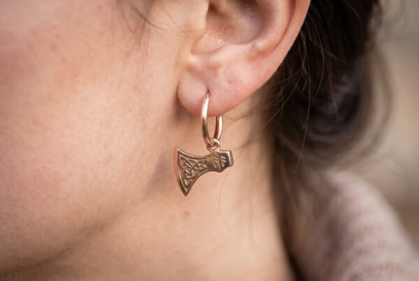 Rose gold tone Viking axe hoop earring with Celtic knot head worn on the ear, shown close up against soft brown hair