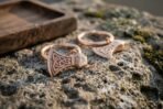 Rose gold tone Viking axe hoop earrings with Celtic knotwork heads lying on mossy stone beside a small wooden tray