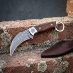 Rosewood Damascus karambit with a rolling wave hawkbill blade, spiral engraved bolster and steel finger ring set on weathered red brick beside a brown leather sheath