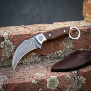 Rosewood Damascus karambit with a rolling wave hawkbill blade, spiral engraved bolster and steel finger ring set on weathered red brick beside a brown leather sheath