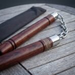 Close up of rosewood nunchaku with knotwork cut collars and steel chain on a grey slatted table, black carry pouch behind