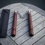 Rosewood nunchaku with polished steel chain laid in a V on a grey wooden table, black cloth pouch to the left, top down view