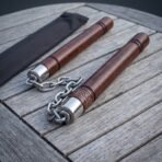Rosewood nunchaku with steel chain and knotwork collars laid side by side on a weathered slatted table beside a black pouch