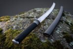 Round brushed steel tsuba and valknut etched blade of a hand forged viking katana resting on a lichen covered boulder beside its charcoal saya
