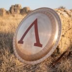 Round Greek hoplite shield with a red painted lambda and engraved meander border leaning on a rock in dry golden grass