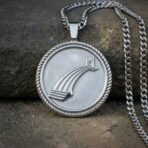 Close up of a round silver tone bifrost pendant with a raised bridge, rope twist border and heavy curb chain on a stone ledge
