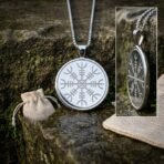 Round silver tone helm of awe pendant with engraved stave on a box chain resting on mossy stone, linen pouch and detail inset