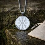 Close up of a round silver tone pendant engraved with the eight armed helm of awe stave on a box chain beside a linen pouch