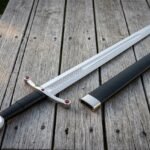 Ruby pommel arming sword in polished 1095 steel with red stone set guard terminals and a black leather grip laid on weathered decking beside its black scabbard