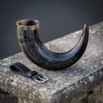 Rune drinking horn in dark polished ox horn with a full Elder Futhark row cut along the curve, a steel rim and a black leather belt loop on stone