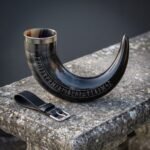 Rune drinking horn in dark polished ox horn with a full Elder Futhark row cut along the curve, a steel rim and a black leather belt loop on stone