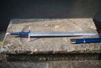 Rune engraved arming sword with a blue leather grip and wheel pommel lying beside a matching blue scabbard on a weathered stone step against a dark wall