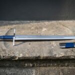 Rune engraved arming sword with a blue leather grip and wheel pommel lying beside a matching blue scabbard on a weathered stone step against a dark wall